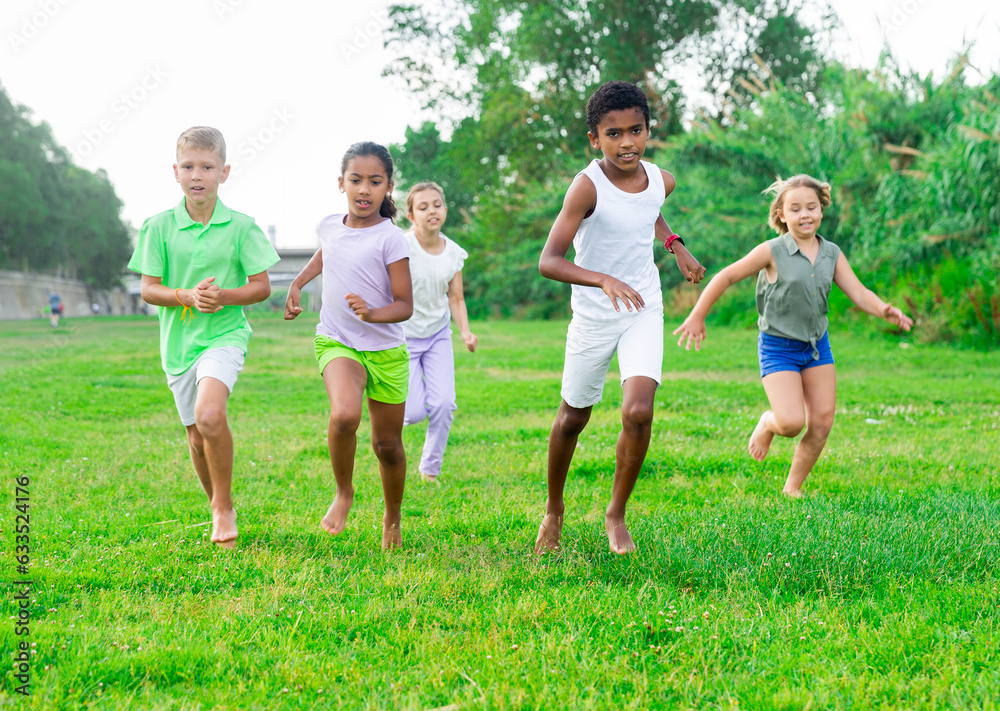 Portrait of international group of happy preteen boys and girls running on green lawn in city park on summer day..