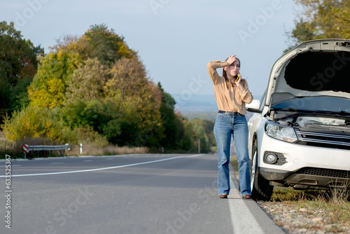 Upset young woman driver talking angrily on cell phone with assistance service near a broken car with open hood while inspecting engine having trouble with her vehicle.