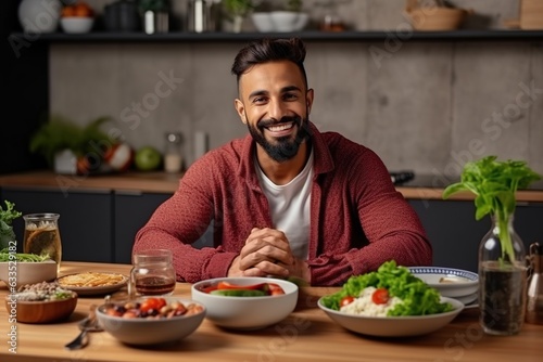 Young fit man having healthy breakfast at home, with a lot of fresh vegetables on kitchen table. Handsome indian man starting day with healthy meal.
