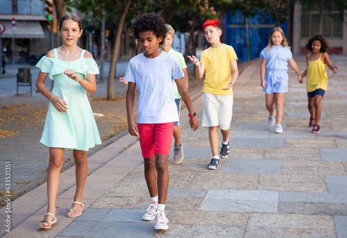 Cute preteen girl talking friendly with her african american friend while walking together along city street on summer day..