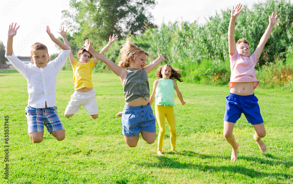 Group of children, boys and girls, jumping together on green grass in a park on sunny day