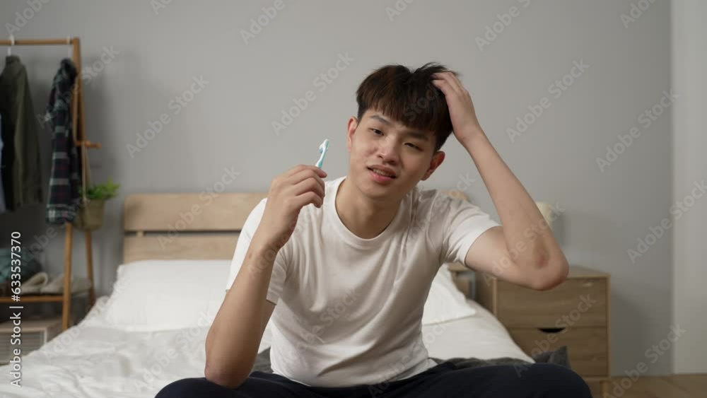 tired asian boy awaking from nodding off while using toothbrush to clean mouth in the morning and scratching head feeling embarrassed in the bedroom at home