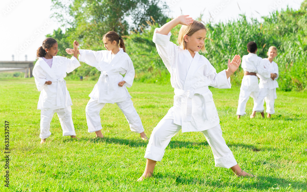 Concentrated tween girl in white kimono exercising new techniques during class taekwondo group outdoors on green lawn on summer day