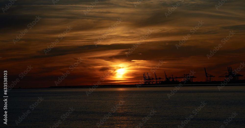 Distant sunset with orange sky and clouds over the sea, with Le Havre port cranes in the background