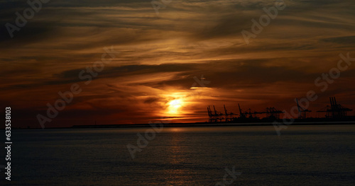 Distant sunset with orange sky and clouds over the sea, with Le Havre port cranes in the background © Sylvain