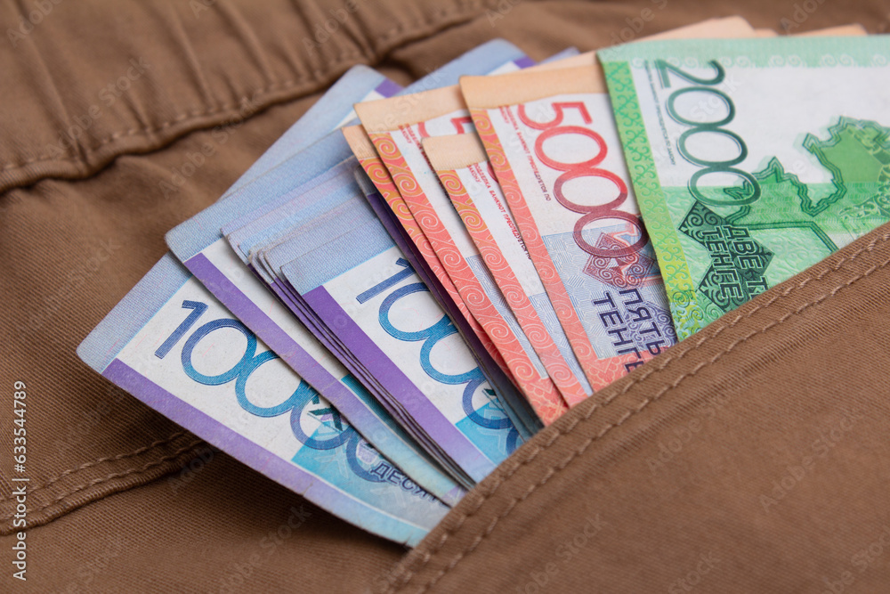 Obraz premium Paper multicolored money of republic of kazakhstan in pocket of brown denim jeans. Textile background. Vacation cash, salary, currency exchange, immigration, travel, trip, bribe, loan. Kazakh tenge