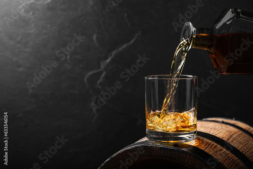 Pouring whiskey into glass from bottle on wooden barrel against grey background, space for text