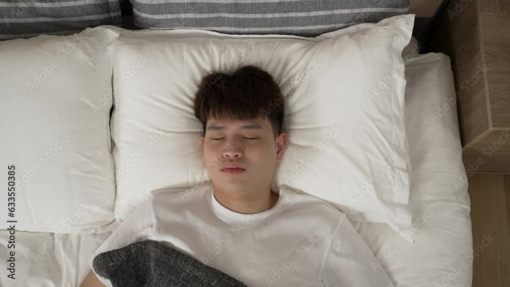top angle shot with zoom in restless asian boy awaking to bad dream is patting his chest and staring upward at camera while breathing heavily in bed at home top angle shot with zoom in restless asian boy awaking to bad dream is patting his chest and staring upward at camera while breathing heavily in bed at home