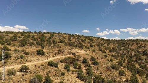 A black Jeep on county dirt road going up the side of a hill on a summer day with white clouds in a blue sky in Arizona.