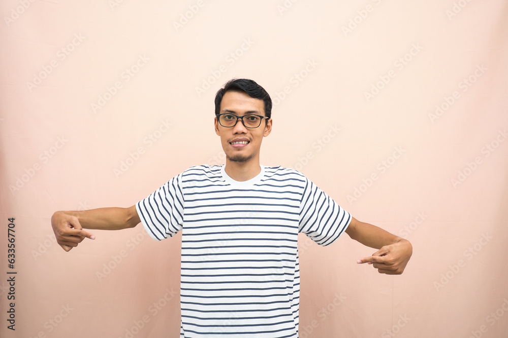 Asian man with glasses wearing casual striped shirt, whispering pose while pointing down. Isolated beige background. Asian man with glasses wearing casual striped shirt, whispering pose while pointing down. Isolated beige background.