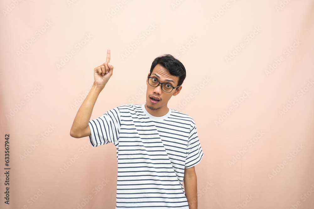 Asian man in glasses wearing casual striped t-shirt, pointing pose above empty space. Isolated beige background. Asian man in glasses wearing casual striped t-shirt, pointing pose above empty space. Isolated beige background.