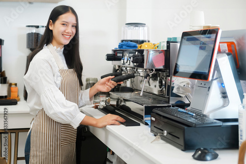 Young cheerful barista wearing apron while preparing coffee at an automatic machine in a modern coffee shop. © ijeab