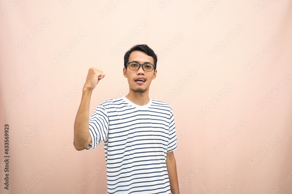 Asian man with glasses wearing casual striped shirt, fist fist pose, indicating strong or ready to fight. Isolated beige background. Asian man with glasses wearing casual striped shirt, fist fist pose, indicating strong or ready to fight. Isolated beige background.