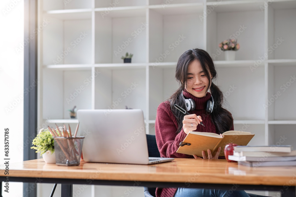 Asian female student studying online using headphones and laptop taking notes in notebook sitting at home table relaxing and changing places to school relax. Asian female student studying online using headphones and laptop taking notes in notebook sitting at home table relaxing and changing places to school relax.