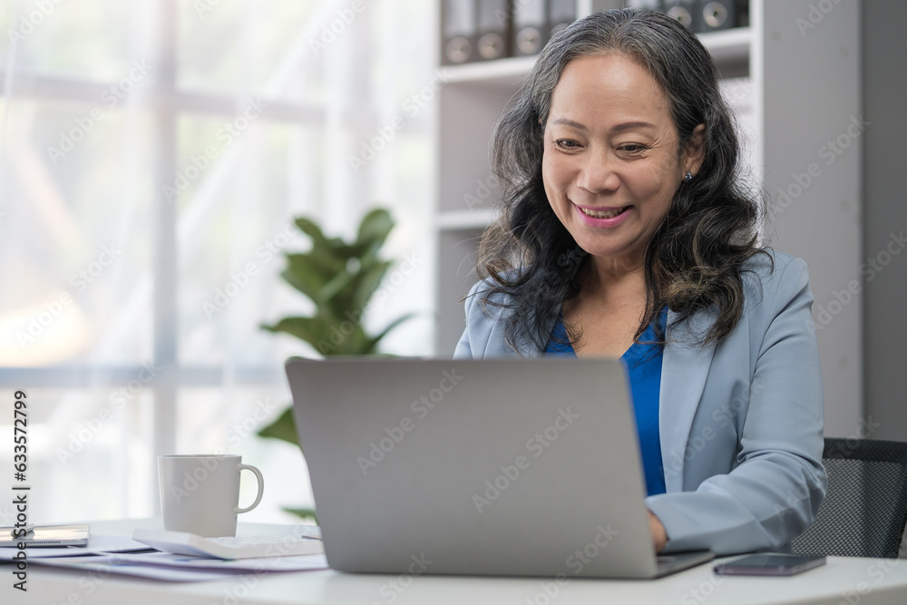 Aged businesswoman reading information on paperwork and using laptop working on desk. Aged businesswoman reading information on paperwork and using laptop working on desk.