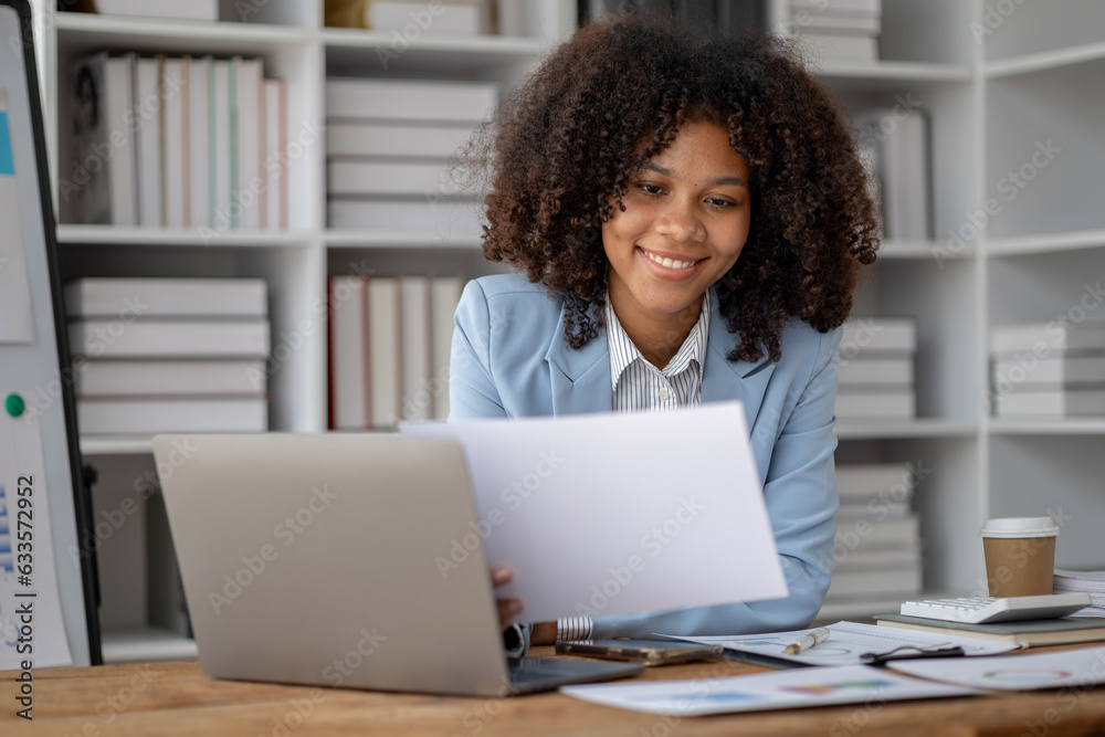 American woman sitting working in a startup company, american businesswoman working in the office, she is looking at financial documents and sales data to summarize before meeting with executives. American woman sitting working in a startup company, american businesswoman working in the office, she is looking at financial documents and sales data to summarize before meeting with executives.