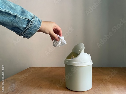 Female hand throwing away a crumpled tissue in small dustbin above wooden table photo