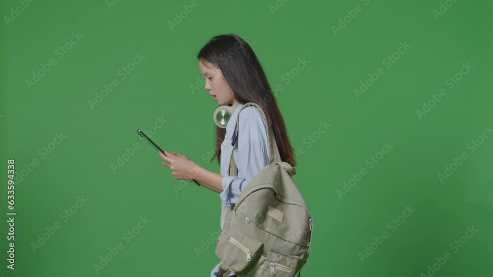Side View Of Asian Teen Girl Student With A Backpack Using A Tablet While Walking In The Green Screen Background Studio
