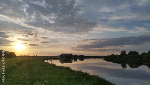 On a summer evening, the sun sinks below the horizon. The colorful cloudy sky and trees are reflected in the quiet river water. The high bank of the river is overgrown with grass. A forest grows