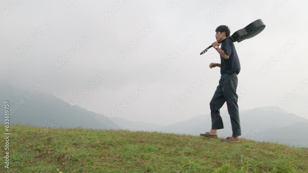 Stylish asian man carrying his guitar on shoulder in mountains. Low angle slow motion view Stylish asian man carrying his guitar on shoulder in mountains. Low angle slow motion view