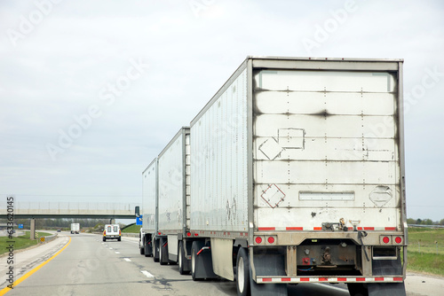 Three trailer road train transporting commercial cargo in USA highway. White vehicle rear cloudy sky