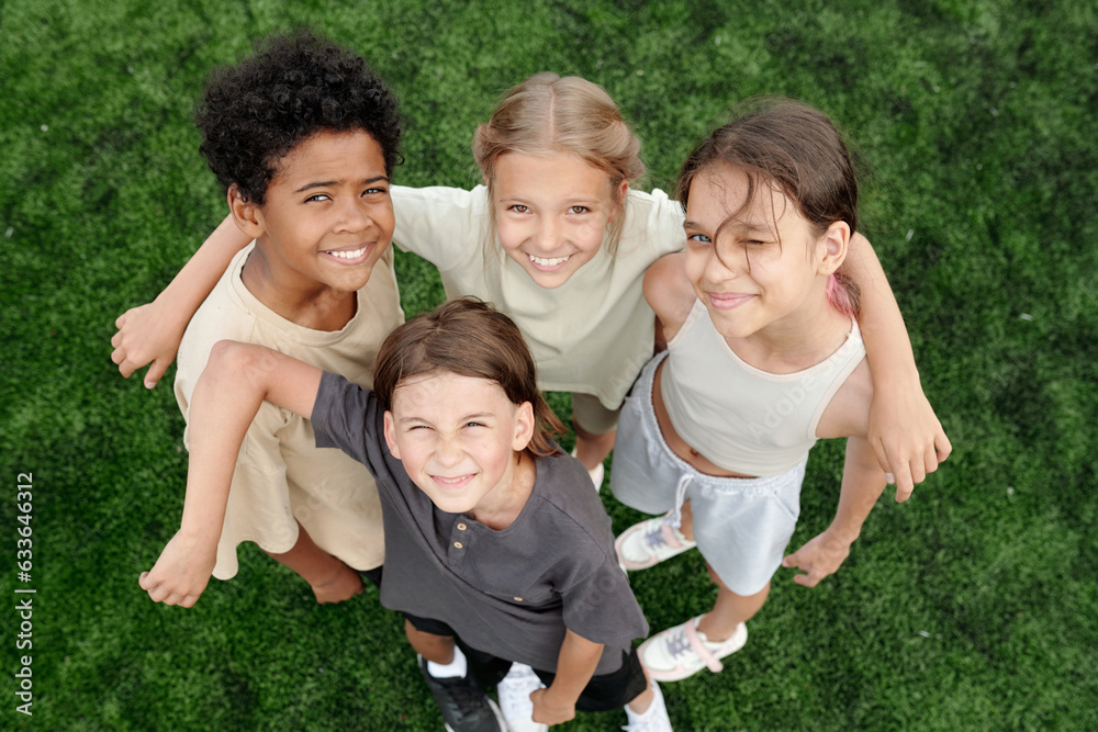 Fototapeta premium Above angle of several happy intercultural children in activewear standing against green lawn or football field and looking at camera with smiles