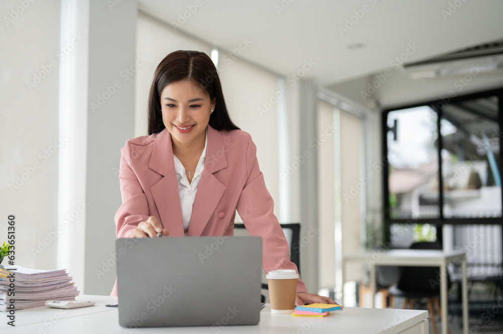 A beautiful Asian businesswoman bending over a table and working on her tasks on her laptop
