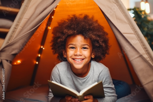 Happy cute little african american kid curly boy reading book. Child reading in tent. photo