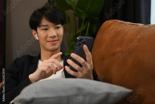 Happy asian man typing text massage on smartphone while resting on couch at home