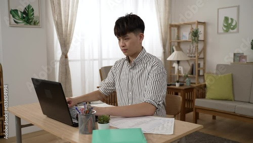 asian businessman doing paperwork with concentration while working from on the laptop in a bright modern living room at home.