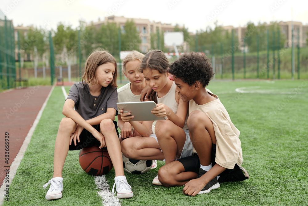 Several intercultural schoolchildren in activewear looking at tablet screen while sitting in front of camera on football field and watching video Several intercultural schoolchildren in activewear looking at tablet screen while sitting in front of camera on football field and watching video