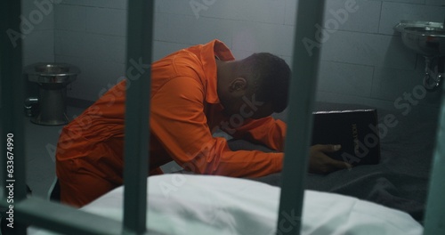 Foto Religious African American prisoner in orange uniform kneels near the bed, prays to God in prison cell holding Bible