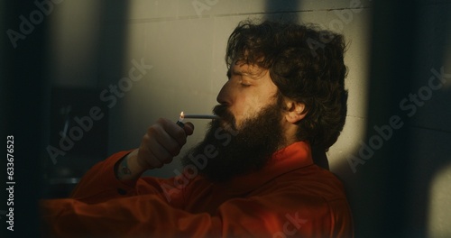 Thoughtful male prisoner in orange uniform sits on the bed, lights cigarette with lighter and smokes in prison cell. Criminal, inmate serves imprisonment term for crime in jail. Detention center.
