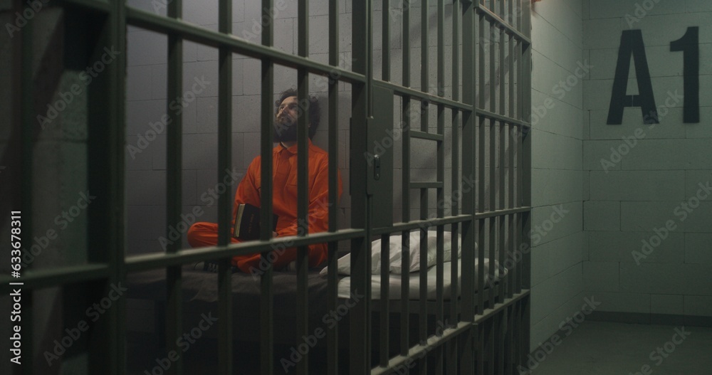 Male prisoner in orange uniform sits on the bed, reads Bible in prison cell. Prison officer walks, watches criminal. Inmate serves imprisonment term in jail. Detention center or correctional facility.