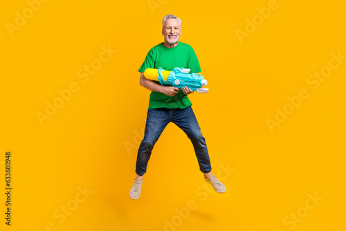Full body portrait of carefree excited aged man jumping arms hold water gun have good mood isolated on yellow color background