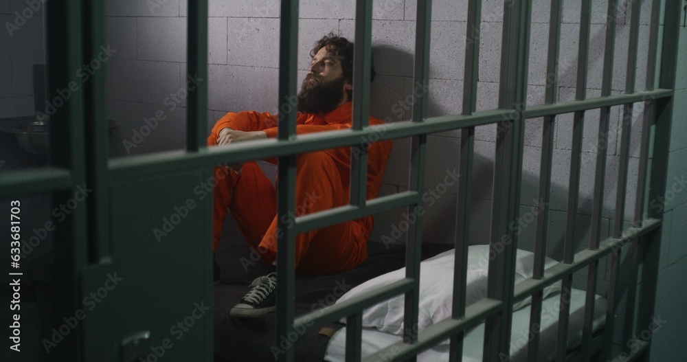 Male prisoner in orange uniform sits on bed, reads Bible, prays, looks at barred window in prison cell. Inmate serves imprisonment term for crime in jail. Correctional facility. Faith in God concept. Male prisoner in orange uniform sits on bed, reads Bible, prays, looks at barred window in prison cell. Inmate serves imprisonment term for crime in jail. Correctional facility. Faith in God concept.