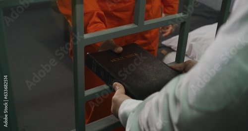 Prison officer gives Bible to male prisoner in orange uniform. Criminal sits on bed in prison cell, reads. Offender serves imprisonment term for crime in jail. Detention center. Faith in God concept.
