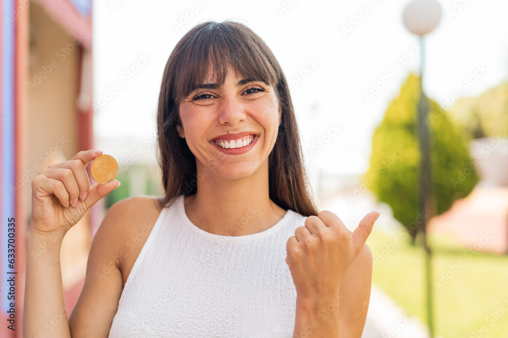 Young woman holding a Bitcoin at outdoors pointing to the side to present a product Young woman holding a Bitcoin at outdoors pointing to the side to present a product