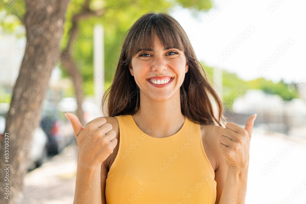 Young woman at outdoors with thumbs up gesture and smiling Young woman at outdoors with thumbs up gesture and smiling