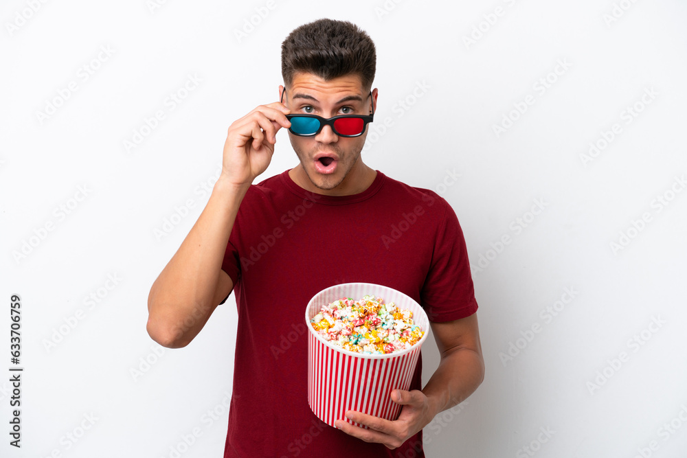 Young caucasian man isolated on white background surprised with 3d glasses and holding a big bucket of popcorns
