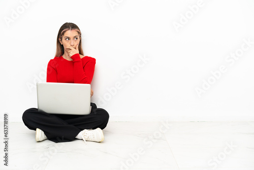 Young caucasian woman with a laptop sitting on the floor having doubts and with confuse face expression
