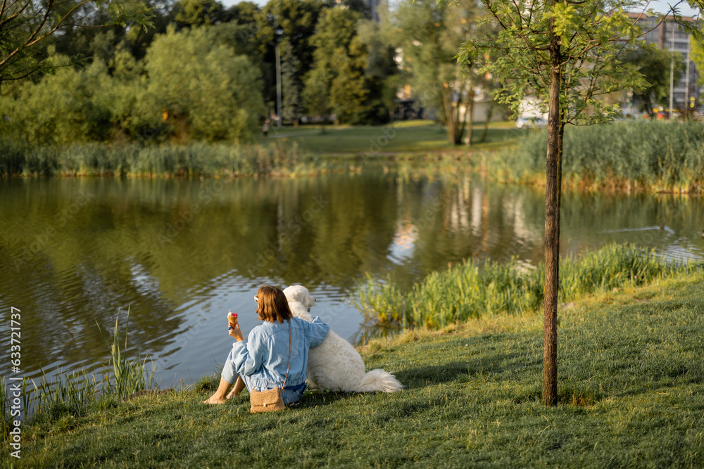Young woman eats ice cream while sitting with her cute white dog near the lake in park. Wide view. Friendship with pets and spending leisure time together Young woman eats ice cream while sitting with her cute white dog near the lake in park. Wide view. Friendship with pets and spending leisure time together