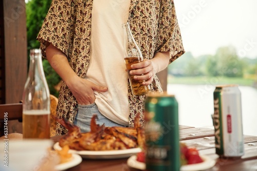 Fototapeta Naklejka Na Ścianę i Meble -  Close up view of woman that is standing by table that is with food, holding beer bottle