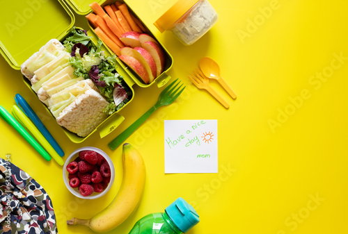 School lunch box with sandwiches, carrot sticks, apple, banana, lettuce, hummus and raspberries. Healthy school lunch concept. Yellow background and note with text - have a nice day mom. Top view 