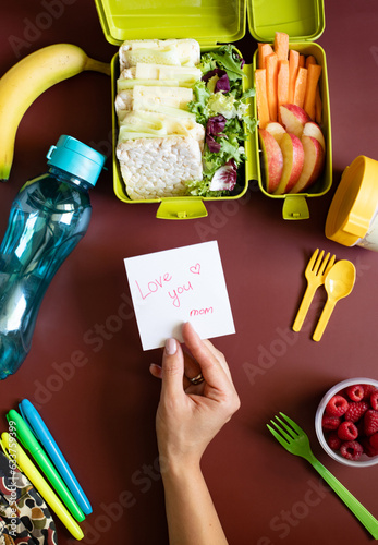 School lunch box with sandwiches, carrot sticks, apple, banana, lettuce, hummus and raspberries. Healthy school lunch concept. Female hand holding a note with text - love you mom. Top view, copy space