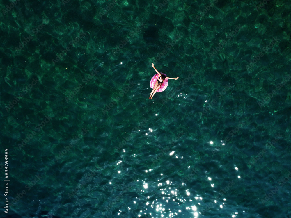 Aerial drone view of young woman with open arms in swimsuit lying and relaxing on big pink donut float swims on water surface with transparent bottom. Summer holiday vacation at exotic resort