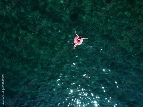 Aerial drone view of young woman with open arms in swimsuit lying and relaxing on big pink donut float swims on water surface with transparent bottom. Summer holiday vacation at exotic resort © Andriy Medvediuk