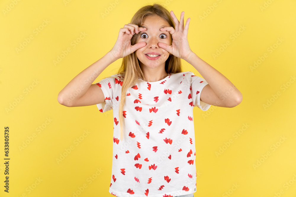 blonde kid girl wearing polka dot shirt over yellow studio background keeping eyes opened to find a success opportunity.