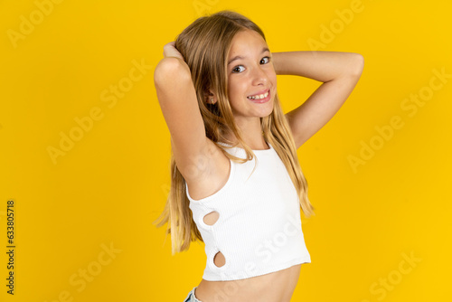 blonde kid girl wearing white T-shirt over yellow studio background stretching arms, relaxed position.