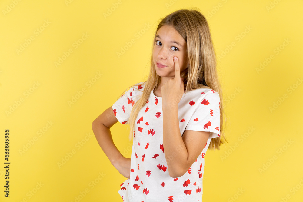 blonde kid girl wearing polka dot shirt over yellow studio background Pointing to the eye watching you gesture, suspicious expression. blonde kid girl wearing polka dot shirt over yellow studio background Pointing to the eye watching you gesture, suspicious expression.
